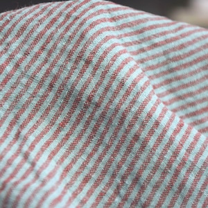 Può includere: Primo piano di un tessuto piegato con un motivo a righe. Il tessuto presenta strisce alternate di blu chiaro e marrone rossiccio. La trama del tessuto è visibile, suggerendo un materiale di lino o cotone.