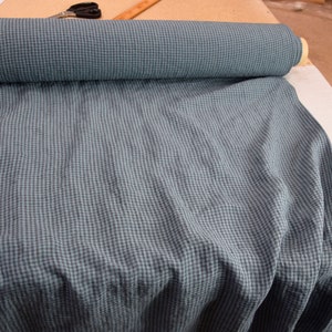 May include: A roll of blue and teal checkered fabric, partially unrolled, revealing the pattern. The fabric is on a white surface, with scissors and a ruler visible in the background. The fabric appears to be linen.