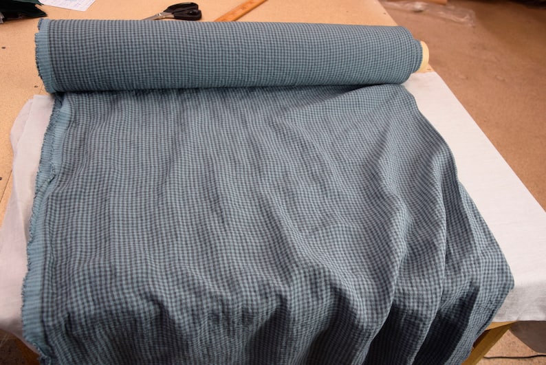 May include: A roll of blue and teal checkered fabric, partially unrolled, revealing the pattern. The fabric is on a white surface, with scissors and a ruler visible in the background. The fabric appears to be linen.