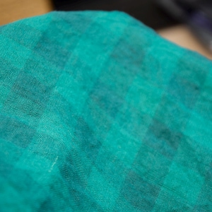 May include: Close-up of a teal fabric with a checkered pattern. The fabric has a soft, textured appearance, with darker teal squares interspersed throughout the lighter teal background. The fabric appears to be lightweight and possibly linen or cotton.