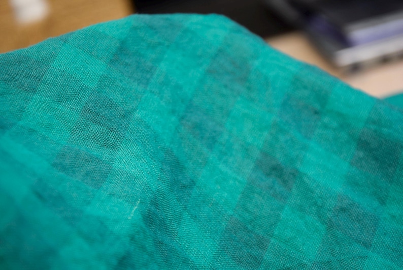 May include: Close-up of a teal fabric with a checkered pattern. The fabric has a soft, textured appearance, with darker teal squares interspersed throughout the lighter teal background. The fabric appears to be lightweight and possibly linen or cotton.