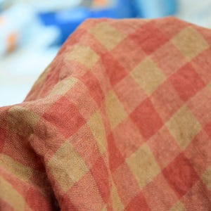 以下が含まれることがあります： 赤とベージュのチェック柄の生地のクローズアップ。 生地は折りたたまれており、質感と色のバリエーションを示しています。 チェック柄のデザインは、さまざまなテキスタイル用途に適したクラシックなパターンです。