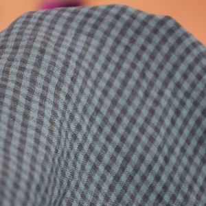 May include: Close-up of a fabric with a checkered pattern. The fabric has a dark gray and teal color scheme, with a repeating square design. The material appears soft and slightly textured, with a subtle sheen. The fabric is likely used for clothing or home decor.