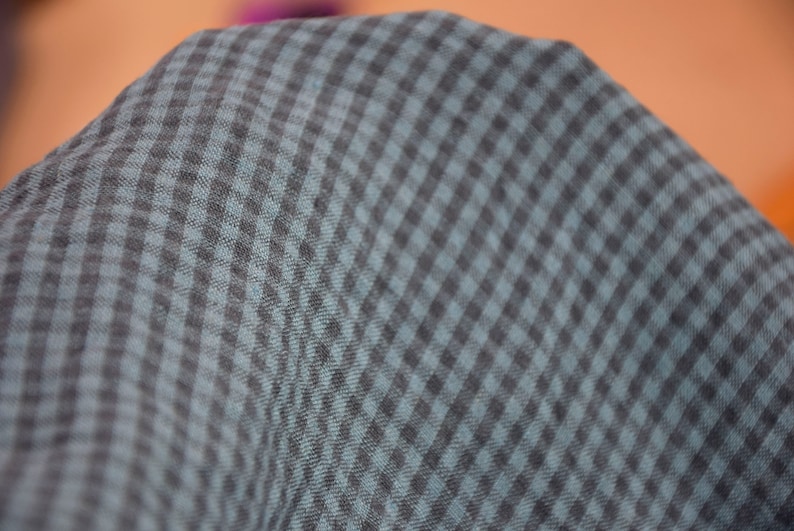 May include: Close-up of a fabric with a checkered pattern. The fabric has a dark gray and teal color scheme, with a repeating square design. The material appears soft and slightly textured, with a subtle sheen. The fabric is likely used for clothing or home decor.