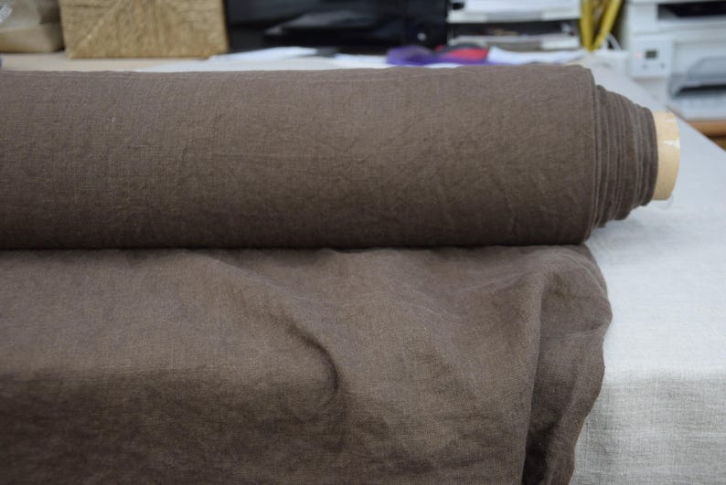 May include: A roll of brown fabric sits atop a flat surface with a portion of the fabric unrolled. The fabric appears to be a linen or similar material, with a slightly textured surface. The color is a muted, earthy brown.