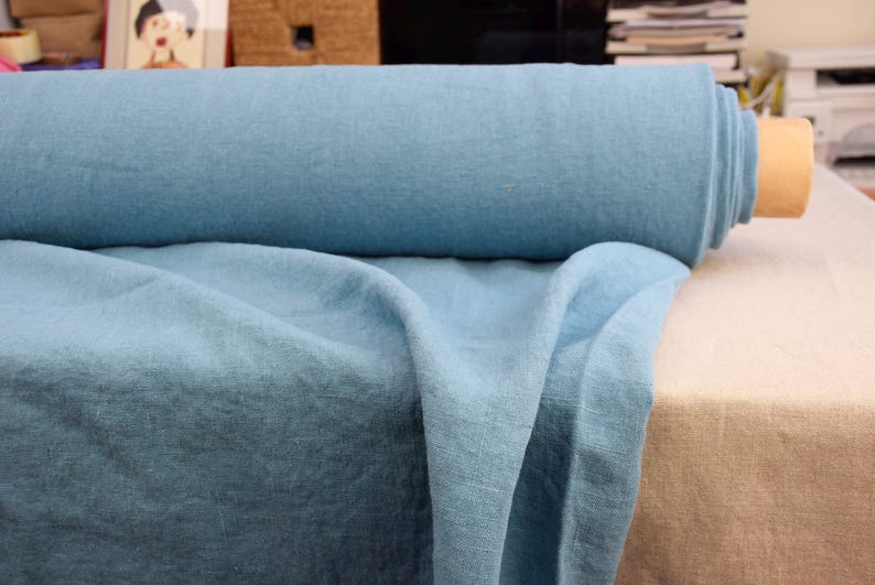 May include: A roll of light blue fabric, possibly linen, is displayed. The fabric is draped over a table, revealing its texture and color. The roll is on a cardboard core. The fabric appears soft and suitable for clothing or home decor.