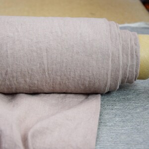 Puede incluir: Un rollo de tela de lino suave de color rosa claro. La tela está tejida con una textura sutil y tiene una sensación natural, ligeramente áspera.
