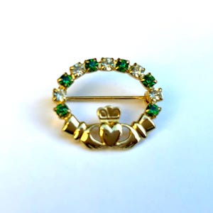 May include: A gold-toned Celtic knot pin with a heart in the center and a crown on top. The knot is surrounded by a circle of green and white rhinestones.