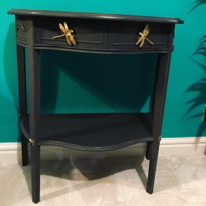 Op de afbeelding: Een donkerblauwe consoletafel met een gebogen bovenblad en twee laden. De laden hebben gouden libellen-grepen. De tafel heeft een onderplank en staat op vier poten.