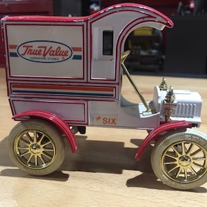May include: A white and red toy delivery truck with a gold-colored steering wheel and wheels. The truck has a blue and red stripe with the words "True Value Hardware Stores" on the side. The truck is labeled with the number "# Six".