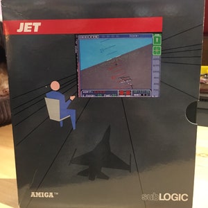 Pode incluir: Uma caixa preta com uma faixa vermelha e a palavra "JET" em letras brancas. A caixa apresenta uma silhueta de um caça e uma captura de tela da interface do jogo. O texto "AMIGA TM" e "subLOGIC" estão impressos na caixa.