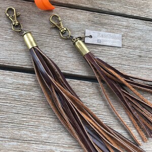 May include: Two leather tassels with bronze-colored hardware. The tassels are made of dark brown and tan leather strips. Each tassel has a bronze-colored metal cap and a lobster claw clasp. The tag reads "RawBoho".