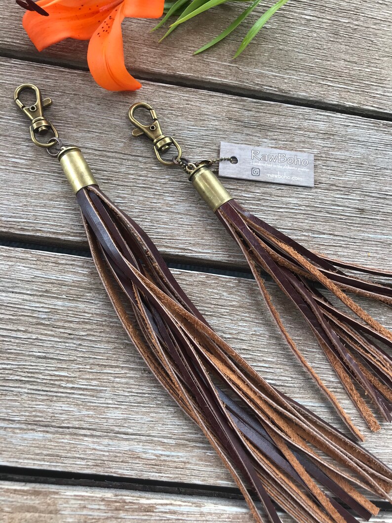 May include: Two leather tassels with bronze-colored hardware. The tassels are made of dark brown and tan leather strips. Each tassel has a bronze-colored metal cap and a lobster claw clasp. The tag reads "RawBoho".