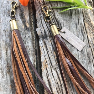 May include: Two leather tassel keychains with brass hardware. The tassels are a mix of dark brown and tan leather strips. The keychains are displayed on a weathered wooden surface, with an orange flower and green leaves in the background.