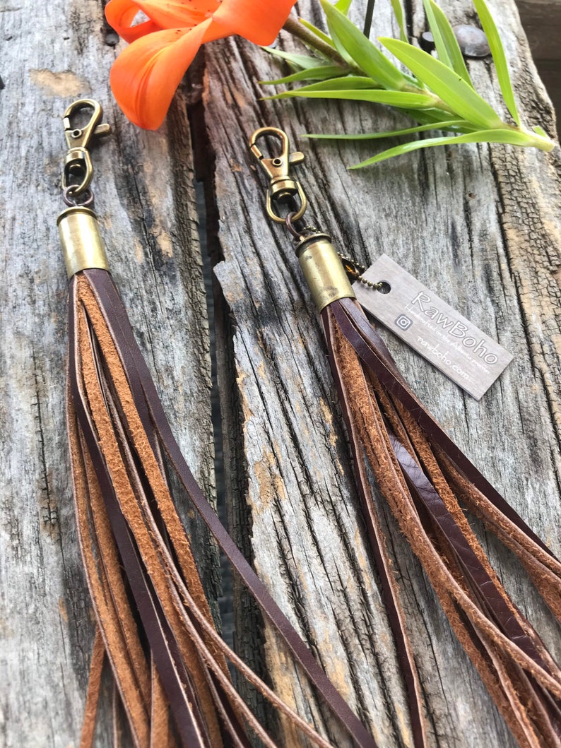 May include: Two leather tassel keychains with brass hardware. The tassels are a mix of dark brown and tan leather strips. The keychains are displayed on a weathered wooden surface, with an orange flower and green leaves in the background.