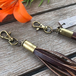 May include: Two brown leather tassels with brass-colored hardware and swivel clasps. The tassels are made of multiple strips of leather. An orange lily flower and green leaves are in the background.