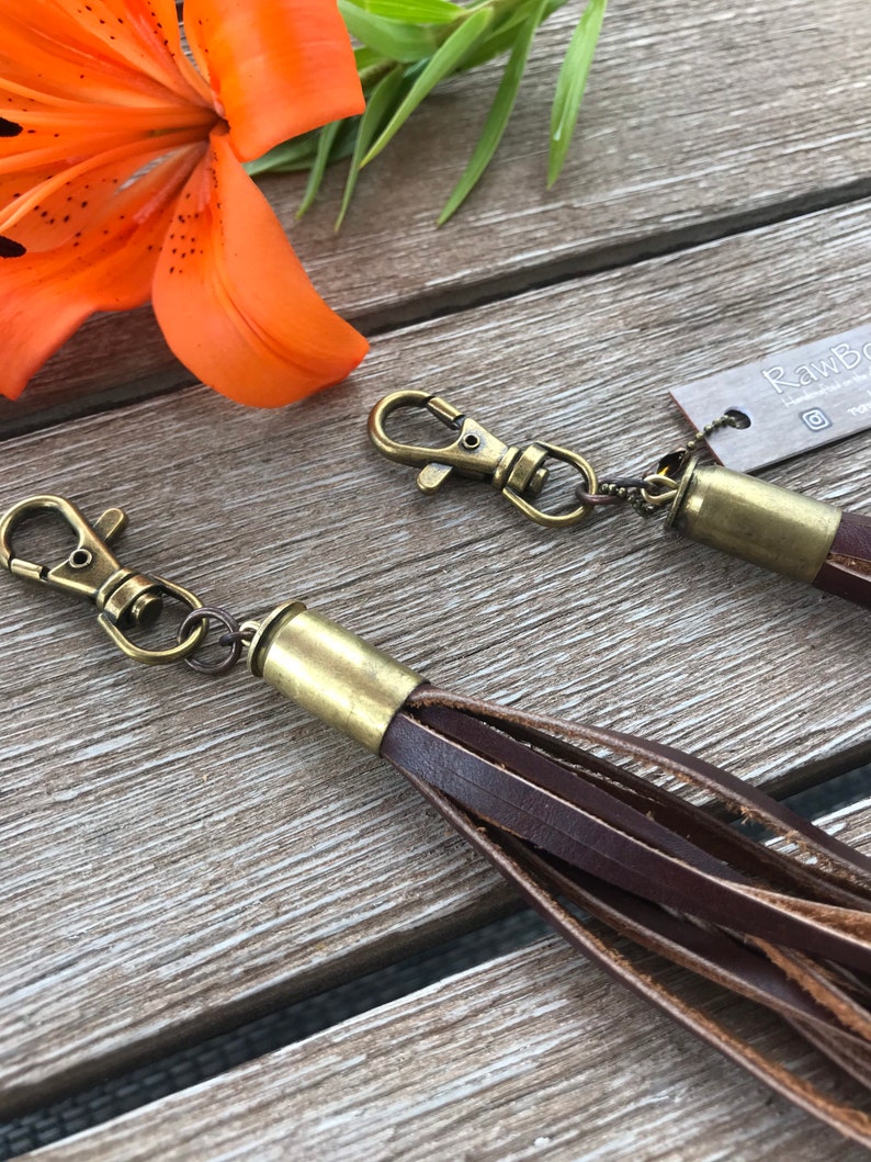 May include: Two brown leather tassels with brass-colored hardware and swivel clasps. The tassels are made of multiple strips of leather. An orange lily flower and green leaves are in the background.