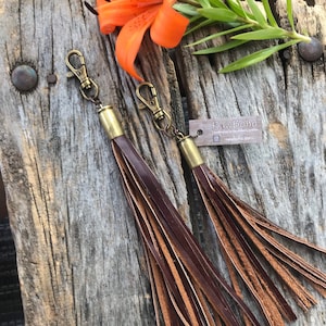 May include: Two leather tassel keychains with brass-colored hardware and lobster claw clasps. The tassels are made of layered brown leather strips. The keychains are displayed on a weathered wooden surface, with an orange flower and green leaves in the background. The tag reads "rawBoho".