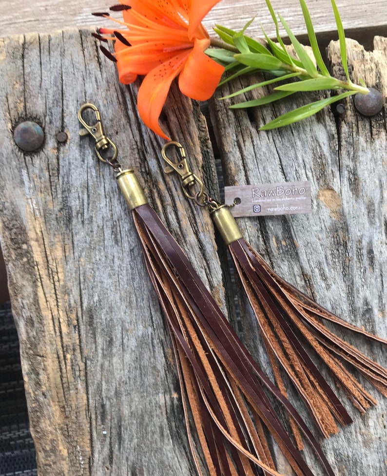 May include: Two leather tassel keychains with brass-colored hardware and lobster claw clasps. The tassels are made of layered brown leather strips. The keychains are displayed on a weathered wooden surface, with an orange flower and green leaves in the background. The tag reads "rawBoho".