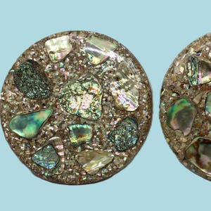 May include: Two round, mosaic-style earrings. Each earring features a speckled, light brown base embedded with iridescent abalone shell pieces in shades of green, blue, and silver. The earrings have a polished finish.