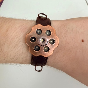 Peut inclure: Une montre de couleur cuivre avec un design de barillet de revolver et un bracelet en cuir marron. Le cadran de la montre porte l'inscription "PYTHON 357 MAG". La montre est fixée au poignet par une boucle.