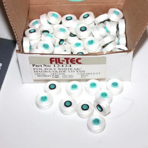 May include: A cardboard box filled with white spools of thread, each with a teal center. Some spools are scattered outside the box. A label on the box reads "FIL-TEC". A document with the text "CORE RETURN" is visible.