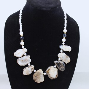 May include: A necklace featuring large, irregularly shaped, light brown and white stones. The stones are interspersed with white and dark blue beads, and small gold-colored accents. The necklace is displayed on a black mannequin.