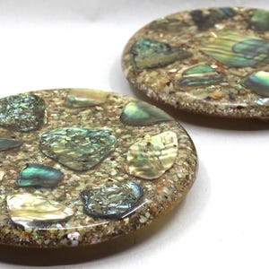 May include: Two oval-shaped decorative coasters. Each coaster features a speckled, light brown base embedded with iridescent abalone shell pieces in shades of green, blue, and gold. The coasters are set against a white background.