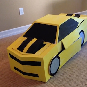 May include: A yellow cardboard car costume with black stripes and a black windshield. The car is made of cardboard and has a simple design.