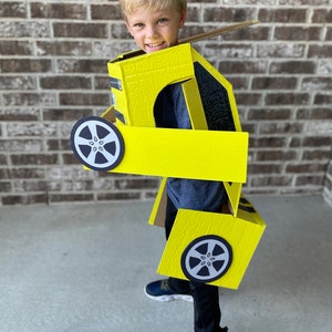 May include: A young boy wearing a yellow cardboard costume of a car. The costume has black wheels and a black stripe. The boy is smiling and looking at the camera.