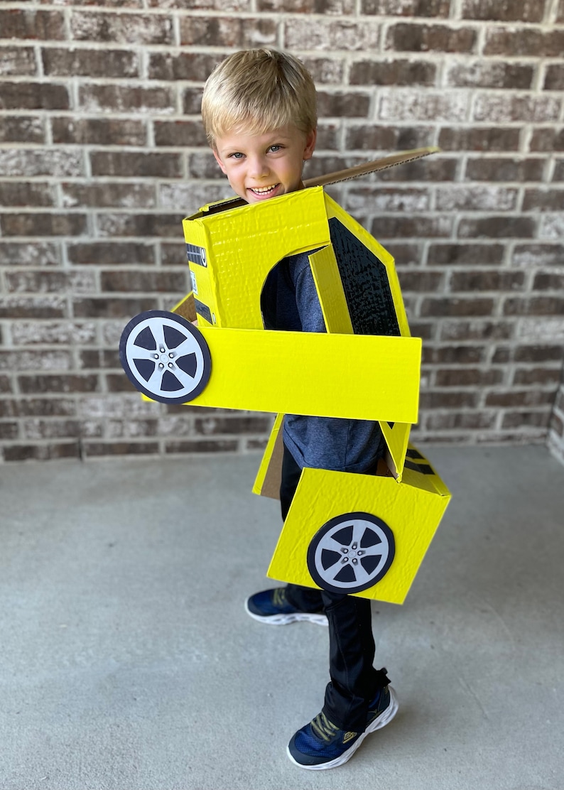 May include: A young boy wearing a yellow cardboard costume of a car. The costume has black wheels and a black stripe. The boy is smiling and looking at the camera.