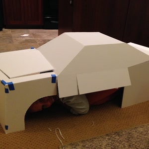 May include: A white cardboard box shaped like a car with a person inside. The box is taped together with blue tape.