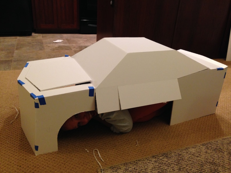 May include: A white cardboard box shaped like a car with a person inside. The box is taped together with blue tape.