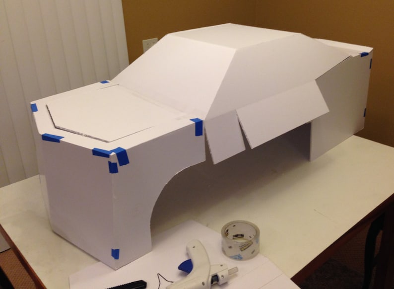 May include: A white cardboard model of a car with blue painter's tape marking the seams. The model is sitting on a white table with a roll of tape and a glue gun.