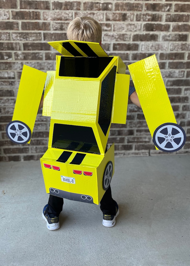 May include: A child wearing a yellow cardboard costume of a Bumblebee Transformer car. The costume has black stripes, a license plate that reads "BMBl-B", and wheels.