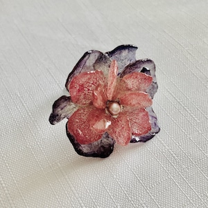 May include: A floral ring with a layered design. The central flower is pink, with a pearl-like centre. The petals are pink and purple, with a silver-coloured band.