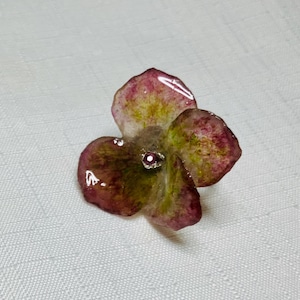 May include: A resin flower brooch with a pink and green color scheme. The flower has a single, small, pink gem in the center.