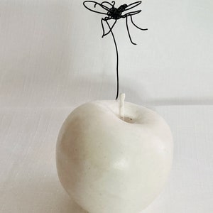 Könnte beinhalten: Eine glatte, cremeweiße apfelförmige Kerze mit einem zentralen weißen Docht. Eine dünne, schwarze Drahtskulptur in Form eines kleinen Insekts ist oben an der Kerze befestigt und ragt in der Nähe des Dochtansatzes nach oben.