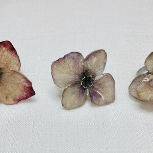 Puede incluir: Tres anillos con forma de flor en tonos beige, morado y burdeos. Cada anillo tiene una pequeña gema redonda morada en el centro. Los anillos están sobre una superficie blanca.