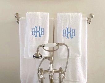 May include: Two white towels with blue embroidered initials "HKH" hanging on a silver towel rack in a bathroom. A silver faucet with a white porcelain handle is in the foreground.