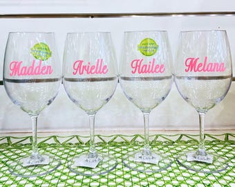 May include: Four clear glass wine glasses arranged in a row, each featuring a unique name printed in bright pink script lettering. The names displayed on the glasses from left to right are Madden, Arielle, Hailee, and Melana. Each glass has a round base.