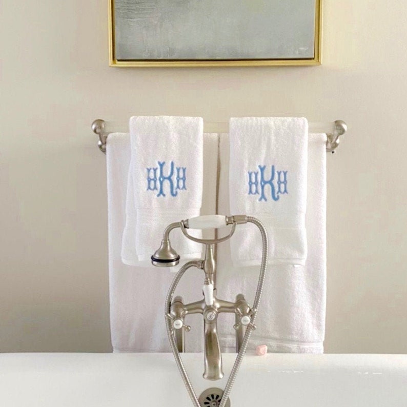 May include: Two white towels with blue embroidered initials "HKH" hanging on a silver towel rack in a bathroom. A silver faucet with a white porcelain handle is in the foreground.