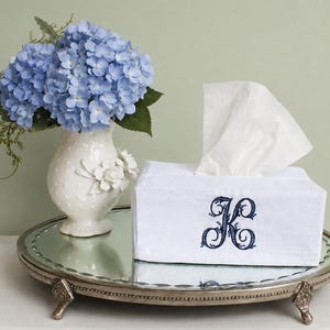 May include: A white rectangular tissue box cover featuring a dark blue embroidered letter K in a decorative script font. The cover is placed on a round mirrored tray next to a white ceramic vase filled with a bouquet of light blue hydrangea flowers.