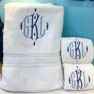 May include: A set of white bath towels with a blue monogram. The large towel is folded and stacked with two smaller towels. The monogram features the letters GKL in a decorative font. The towels have a soft, plush texture.