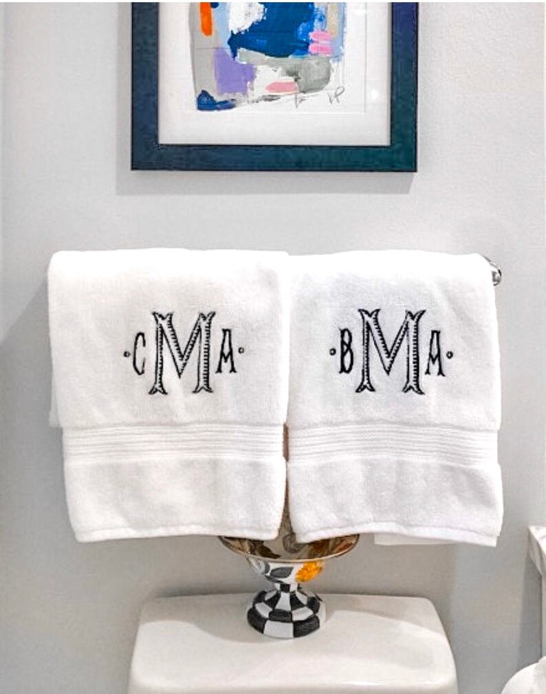 May include: Two white bath towels with black embroidered monograms. The towels are hanging on a towel rack in a bathroom. The monogram on the left towel is "CMA" and the monogram on the right towel is "BMA".
