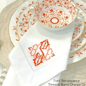 May include: A white linen napkin featuring a monogram with the letter S in burnt orange and the letters B and E in salmon, embroidered in Renaissance font. The napkin rests on white plates with orange floral patterns and gold trim.