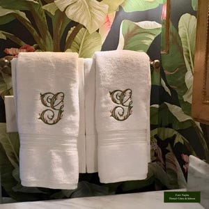 May include: Two white terry cloth hand towels hanging on a metal bar. Each towel features an embroidered monogram of the letter G in a decorative script style, stitched with green and salmon-colored thread. The towels have a ribbed border detail.