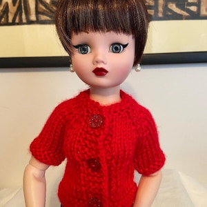 May include: A vintage doll with short brown hair, wearing a red knitted cardigan with three glittery buttons. The doll has blue eyes, red lipstick, and pearl earrings. The doll is wearing black pants.