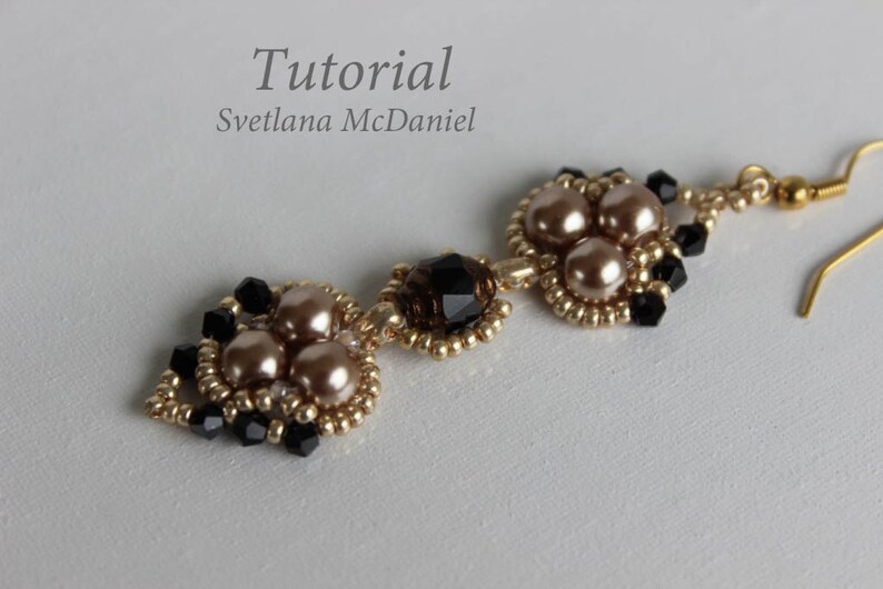 May include: Gold tone earrings with black and beige beads. The earrings feature a cluster of three beige beads surrounded by black beads. The earrings are connected by a gold tone chain with a single black bead.