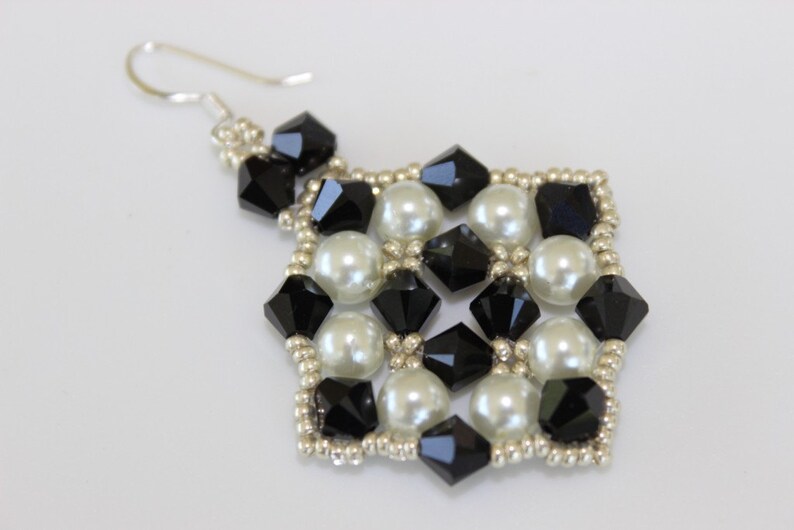 May include: A single beaded earring with a silver hook. The earring features a square design with alternating black faceted beads and white, round, pearl-like beads. The edges are lined with small silver beads.